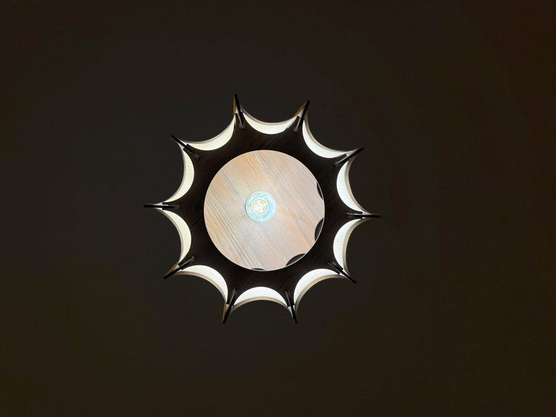 May include: Overhead view of a modern, sunburst-shaped light fixture. The fixture has a dark, spiky outer frame with white interior accents. The center features a light-colored wooden surface with a visible lightbulb.