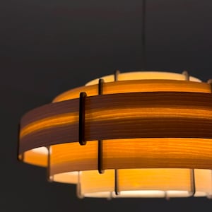 Puede incluir: Una l&aacute;mpara colgante de madera en capas. El dise&ntilde;o presenta m&uacute;ltiples niveles circulares de madera clara, unidos por soportes verticales. La luz emite una iluminaci&oacute;n suave y difusa, creando un ambiente acogedor. La l&aacute;mpara cuelga de un solo cable.