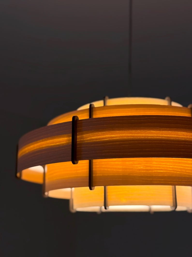 Puede incluir: Una l&aacute;mpara colgante de madera en capas. El dise&ntilde;o presenta m&uacute;ltiples niveles circulares de madera clara, unidos por soportes verticales. La luz emite una iluminaci&oacute;n suave y difusa, creando un ambiente acogedor. La l&aacute;mpara cuelga de un solo cable.