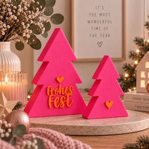 Könnte beinhalten: Zwei leuchtend pinke, dreieckige Weihnachtsbaum-Dekorationen. Der größere Baum zeigt ein orangefarbenes Herz und den orangefarbenen Schriftzug "Frohes Fest" in Schreibschrift. Der kleinere Baum hat ein einzelnes orangefarbenes Herz.