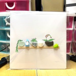 May include: A white card with a shelf design, showcasing miniature plant illustrations. The shelf displays a trailing green plant, a blue gnome, a cactus, a flowering plant, and a green, spiky creature. The card is encased in a clear plastic sleeve.