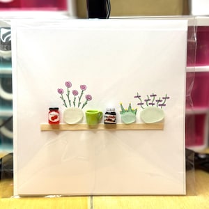 May include: A white card featuring a miniature shelf with tiny decorative items. These include a jar of jam, a green mug, a jar of Nutella, and small plants. Above the shelf are illustrations of flowers and plants.