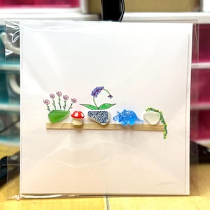 May include: A white card featuring a shelf with colorful glass art. The art includes a green plant, a red mushroom, a purple flower, a blue dinosaur, and a white heart-shaped piece with a green vine.