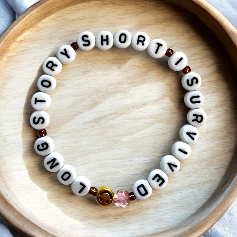 Pu&ograve; includere: Collana di perline bianche con le parole "Long Story Short I Survived" scritte. La collana ha un ciondolo a forma di smiley dorato e un ciondolo a forma di cuore rosa.