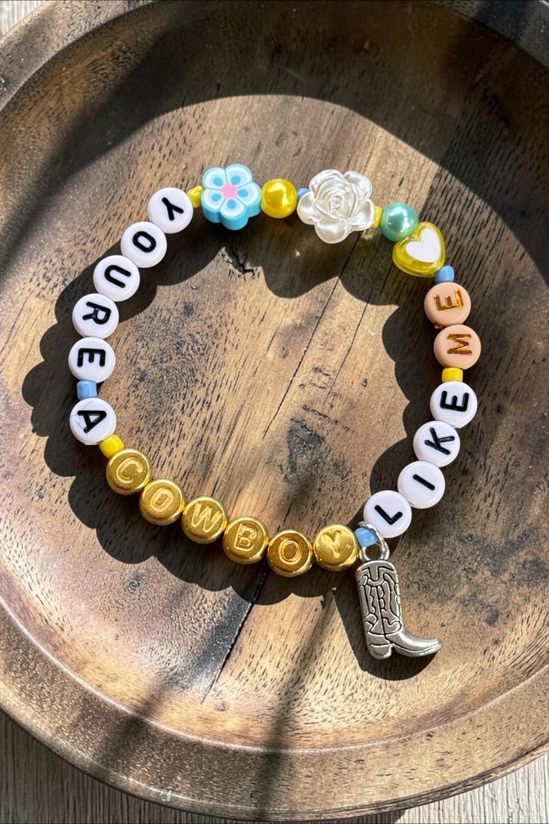 May include: A beaded bracelet featuring white letter beads spelling YOU ARE A COWBOY LIKE ME, interspersed with gold, yellow, and blue beads. The design includes a silver-toned cowboy boot charm, a white flower bead, a blue flower bead, and a yellow heart.