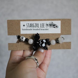 May include: A beaded bracelet featuring a central black bat-shaped charm, flanked by white, black, and translucent beads. The bracelet is attached to a brown cardboard display card labeled STARGIRL LEE handmade beaded bracelet with a fish icon.