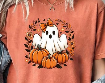 May include: A burnt orange short-sleeve t-shirt featuring a central graphic of a white ghost wearing an orange bow, surrounded by three orange pumpkins and dark foliage. The shirt is worn with a light blue denim jacket draped over the shoulders.