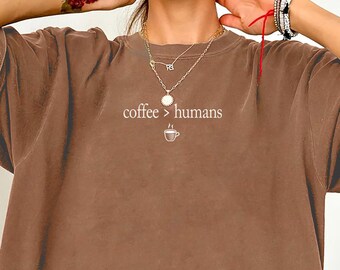 May include: A brown short-sleeve t-shirt featuring the white text "coffee > humans" printed on the chest, accompanied by a small graphic of a coffee cup. The shirt has a relaxed, loose fit with a crew neckline and a solid, matte brown color.