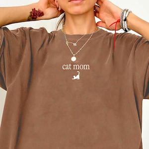 May include: Espresso-colored t-shirt with the text "cat mom" and a cat silhouette. The shirt has a relaxed fit and short sleeves. The fabric appears soft and slightly faded.