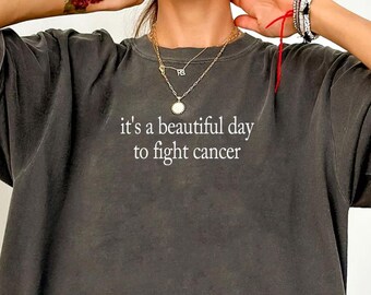 May include: A charcoal grey short-sleeve t-shirt with a washed-out, vintage-style finish. The center of the chest features the white text "it's a beautiful day to fight cancer" printed in a simple, clean, sans-serif font across the front.