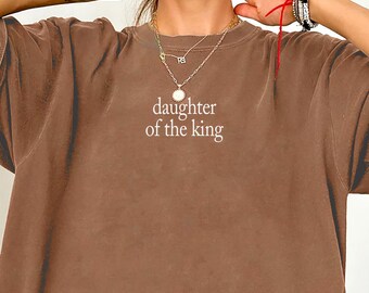 May include: An espresso brown short-sleeve t-shirt featuring the text "daughter of the king" printed in a light cream color on the center of the chest. The shirt has a crew neckline and a loose, relaxed fit with short sleeves and a straight hem.
