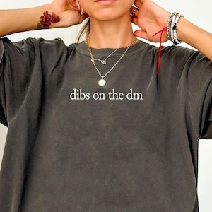 May include: A dark gray oversized t-shirt with the phrase "dibs on the dm" printed in white. The shirt has short sleeves and a relaxed fit, perfect for casual wear. The word "Pepper" is written in cursive in the bottom left corner.