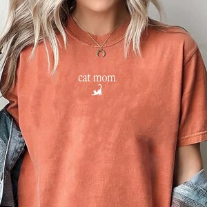 May include: Orange-red t-shirt with the text "cat mom" and a small white cat illustration. The shirt has short sleeves and a relaxed fit. A light blue denim jacket is worn over the shirt.