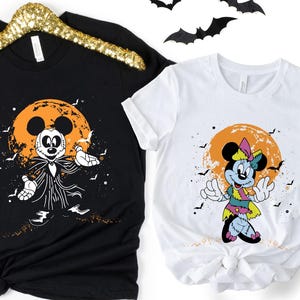 May include: Two black and white t-shirts with a Halloween theme. The shirt on the left features Mickey Mouse dressed as a zombie with a jack-o-lantern moon in the background. The shirt on the right features Minnie Mouse dressed as a zombie with a jack-o-lantern moon in the background.