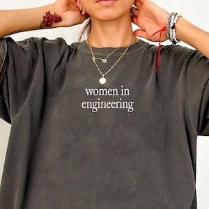 May include: Dark grey t-shirt featuring the white text "women in engineering". The shirt has a loose fit and short sleeves. The word "Pepper" is written in cursive at the bottom left corner.