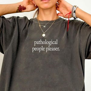 May include: Dark gray t-shirt with the text "pathological people pleaser." The shirt has short sleeves and a relaxed fit. The word "Pepper" is written in cursive in the bottom left corner.