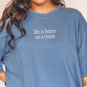 May include: Blue jean-colored t-shirt with the text "life is better on a cruise" in white. The shirt has short sleeves and a relaxed fit. The words "Blue jean" are written in cursive at the bottom of the image.