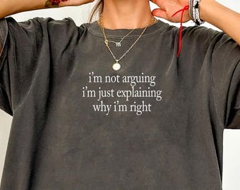 May include: A charcoal gray short-sleeve t-shirt with a crew neckline. The center of the chest features white text that reads, I'm not arguing I'm just explaining why I'm right. The fabric has a washed, vintage-style appearance with a relaxed fit.