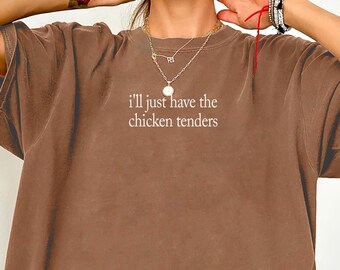 May include: An espresso brown short-sleeve t-shirt featuring the white text "i'll just have the chicken tenders" printed in a small, centered font on the upper chest area. The garment has a crew neckline and a loose, relaxed fit.