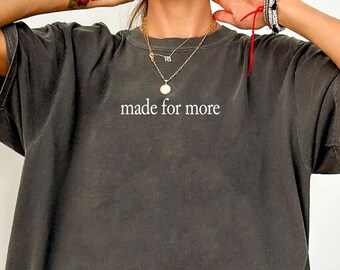 May include: A charcoal gray oversized t-shirt featuring the text "made for more" printed in white on the center chest. The garment has a crew neckline, short sleeves, and a relaxed fit, showcasing a washed-out, vintage-inspired fabric texture.