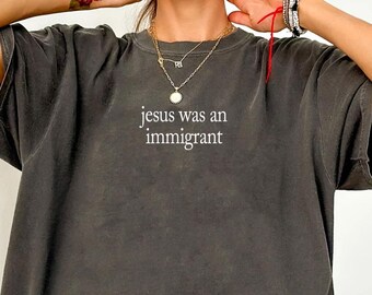 May include: A charcoal grey oversized t-shirt with short sleeves and a crew neckline. The center of the chest features the white text "jesus was an immigrant" printed in a simple, clean, sans-serif font. The fabric has a washed, vintage look.