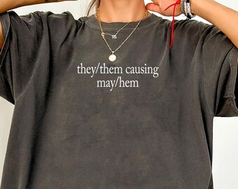 May include: A charcoal grey oversized t-shirt featuring the white text "they/them causing may/hem" printed on the chest. The garment has a crew neckline, short sleeves, and a relaxed fit, showcasing a washed-out, vintage-inspired fabric texture.