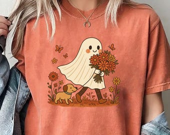 May include: A burnt orange short-sleeve t-shirt featuring a graphic print of a white ghost holding a bouquet of orange flowers, accompanied by a small brown dog, butterflies, and floral elements. The shirt is worn with a blue denim jacket.