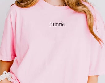 May include: A light pink short-sleeved t-shirt featuring the word auntie printed in a small black serif font on the upper center chest area. The garment has a crew neckline and a relaxed fit, providing a simple and clean aesthetic for daily wear.