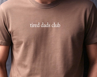 May include: A brown short-sleeved t-shirt featuring the text "tired dads club" printed in white across the chest. The garment has a crew neckline and a standard fit, providing a simple and clean look for casual wear in various settings.