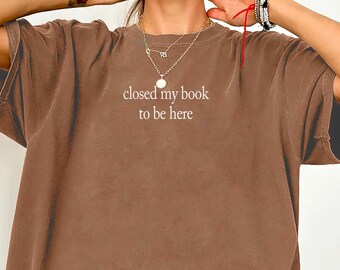 May include: An oversized espresso brown t-shirt featuring the white text "closed my book to be here" printed on the center chest. The garment has a crew neckline, short sleeves, and a relaxed fit, showcasing a solid, muted earth-tone color.