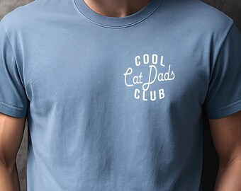 May include: A dusty blue short-sleeved crew neck t-shirt featuring white text on the left chest that reads COOL Cat Dads CLUB in a mix of block and script lettering. The shirt is made of a solid-colored fabric and has a standard fit.