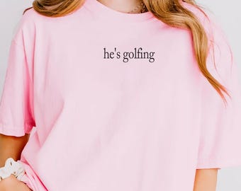 May include: A light pink short-sleeved t-shirt featuring the text he's golfing printed in a small black serif font on the upper left chest area. The garment has a crew neckline and a loose, relaxed fit, paired with blue denim jeans.