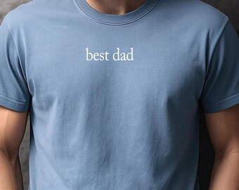 May include: A dusty blue short-sleeved crew neck t-shirt featuring the text "best dad" printed in white across the chest. The shirt is made of a solid-colored fabric and has a standard fit with a ribbed neckline and hemmed sleeves.