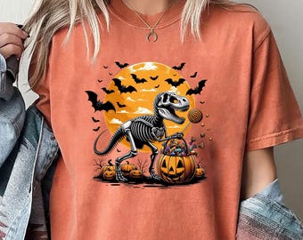 May include: A burnt orange crew neck t-shirt featuring a graphic of a dinosaur skeleton holding a jack-o'-lantern bucket filled with candy. The design includes a large yellow moon, flying bats, and several pumpkins on the ground, all set against the solid orange fabric of the shirt.