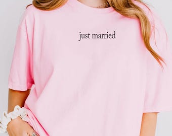 May include: A light pink short-sleeved t-shirt featuring the text "just married" printed in a small, black, serif font on the upper left chest area. The shirt has a crew neckline and a loose, relaxed fit, paired with blue denim jeans.