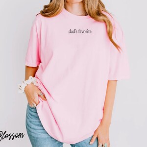 May include: A light pink t-shirt with the text "dad's favorite" printed on the front. The shirt is a soft, comfortable material. The word "Blossom" is written in cursive in the bottom left corner.
