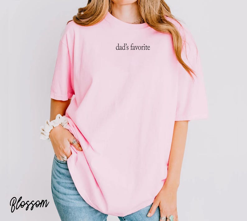 May include: A light pink t-shirt with the text "dad's favorite" printed on the front. The shirt is a soft, comfortable material. The word "Blossom" is written in cursive in the bottom left corner.