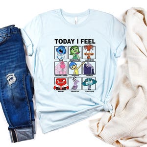 Pu&ograve; includere: Maglietta blu chiaro con una grafica dei personaggi del film Inside Out. La grafica &egrave; una griglia di nove quadrati, ognuno con un personaggio e un'emozione diversi. Il testo "TODAY I FEEL" &egrave; sopra la griglia.
