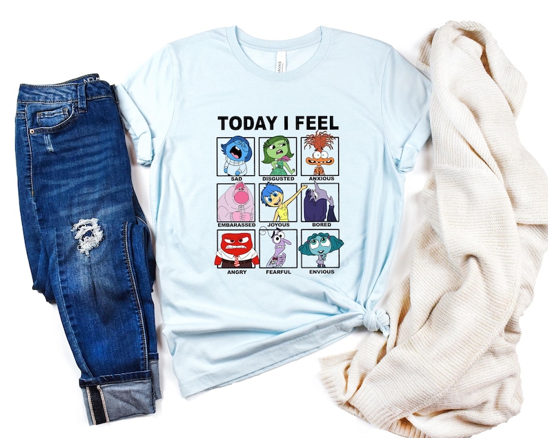 Pu&ograve; includere: Maglietta blu chiaro con una grafica dei personaggi del film Inside Out. La grafica &egrave; una griglia di nove quadrati, ognuno con un personaggio e un'emozione diversi. Il testo "TODAY I FEEL" &egrave; sopra la griglia.