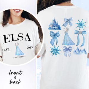 May include: White t-shirt featuring the name "ELSA" in black, with "EST. 2013 ARENDELLE" below a cartoon image of a princess. The back of the shirt displays blue bows, snowflakes, a castle, and a princess.