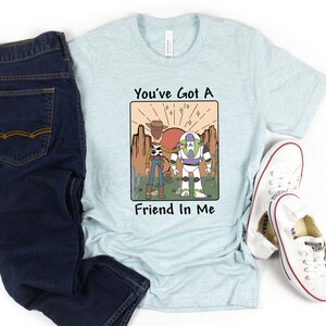 May include: Light blue t-shirt with a graphic featuring Woody and Buzz Lightyear from Toy Story, with the text "You've Got A Friend In Me." The shirt is paired with dark blue jeans and white sneakers.
