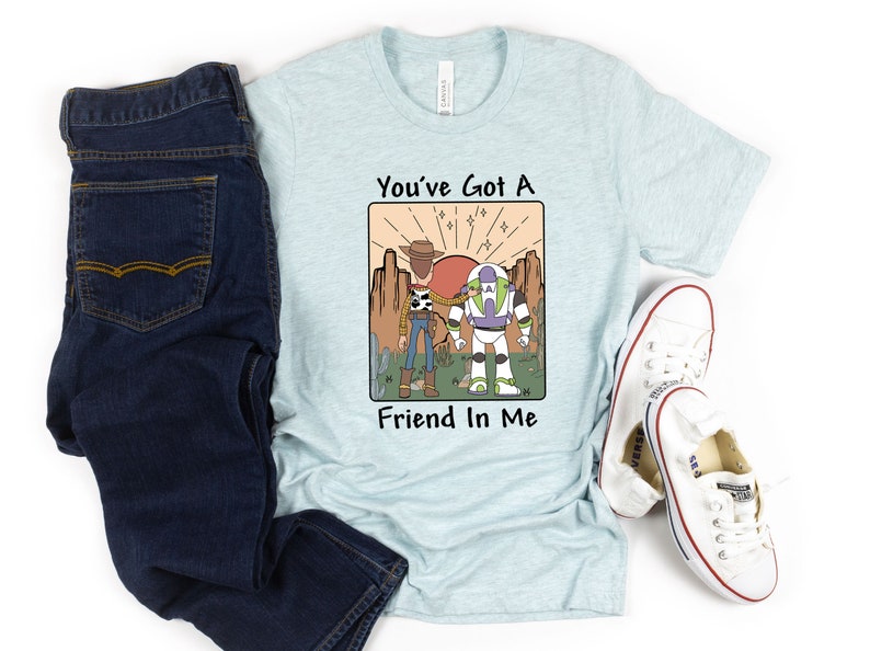 May include: Light blue t-shirt with a graphic featuring Woody and Buzz Lightyear from Toy Story, with the text "You've Got A Friend In Me." The shirt is paired with dark blue jeans and white sneakers.