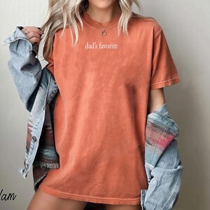 May include: Orange-red t-shirt with the text "dad's favorite" in white. The shirt is worn with a denim jacket. The image has a warm, casual aesthetic.
