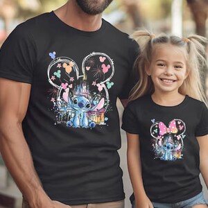 K&ouml;nnte beinhalten: Schwarze T-Shirts mit einer farbenfrohen Grafik der Figur Stitch aus Lilo & Stitch. Das Design umfasst ein Schloss, Feuerwerk und Mickey Mouse Ohren. Die Shirts werden von einem Mann und einem jungen M&auml;dchen getragen.