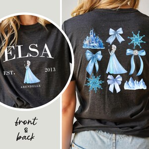 May include: Dark gray t-shirt with a front graphic of "ELSA" and a princess illustration. The back features a castle, bows, snowflakes, and a princess in a blue dress. The text "EST. 2013 ARENDELLE" is also visible.