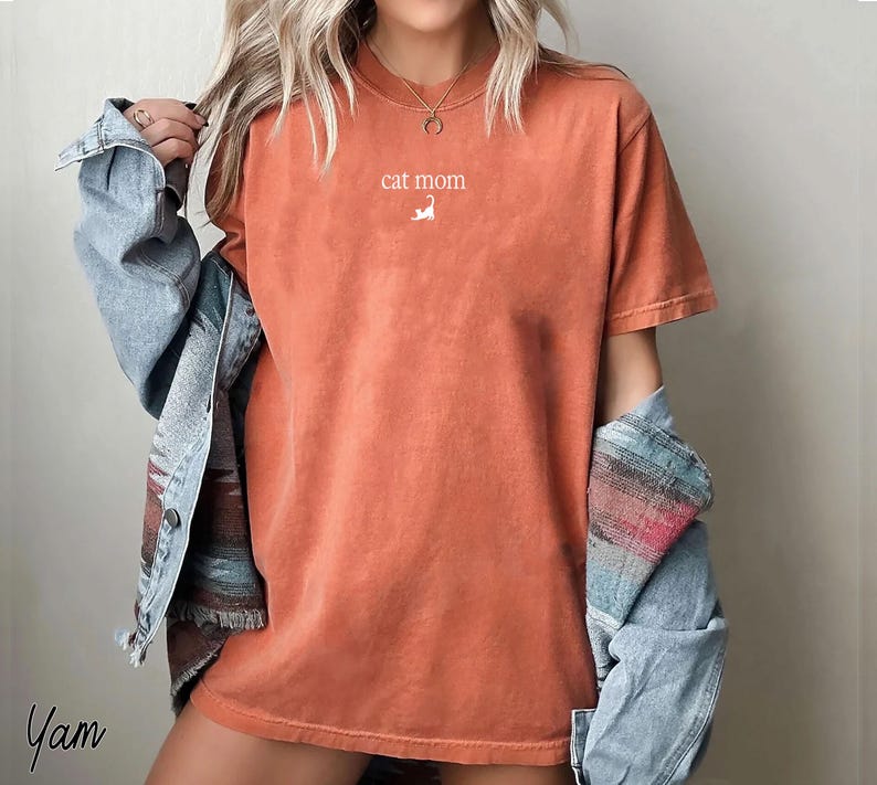 May include: Orange-red t-shirt with the text "cat mom" and a small white cat illustration. The shirt has short sleeves and a relaxed fit. A light blue denim jacket is worn over the shirt.
