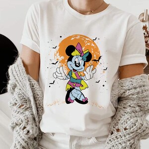 May include: White t-shirt featuring a graphic of a zombie Mickey Mouse character with a pink bow and a green and blue dress. The character is standing in front of a yellow and orange moon with bats flying around it.