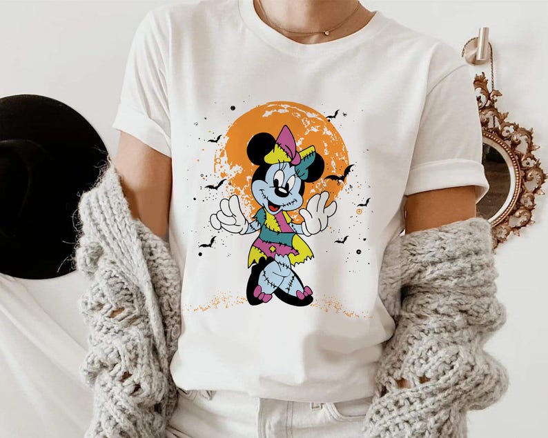 May include: White t-shirt featuring a graphic of a zombie Mickey Mouse character with a pink bow and a green and blue dress. The character is standing in front of a yellow and orange moon with bats flying around it.