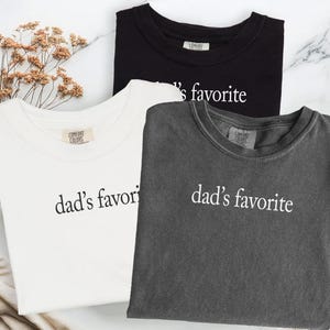 May include: Three t-shirts in black, white, and gray, each with the phrase "dad's favorite" printed in white. The shirts are displayed on a marble surface, with dried flowers in the background.