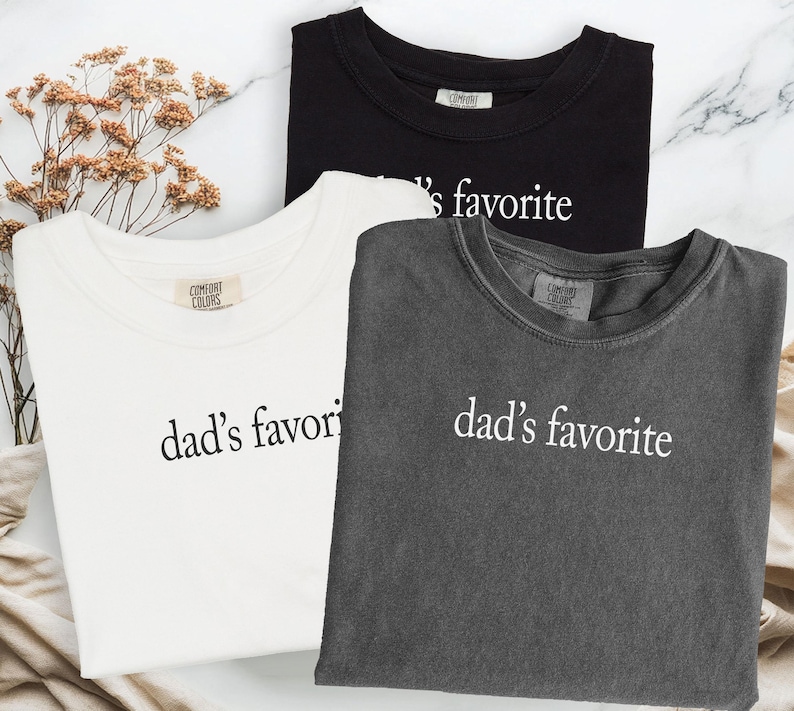 May include: Three t-shirts in black, white, and gray, each with the phrase "dad's favorite" printed in white. The shirts are displayed on a marble surface, with dried flowers in the background.